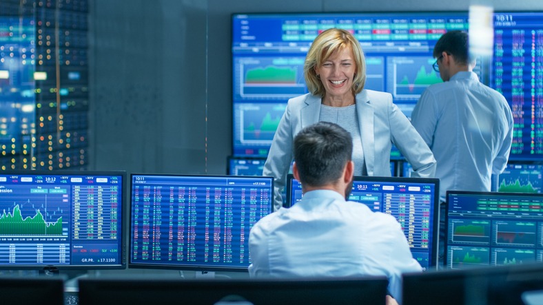 In the Stock Market Selling/ Buying Department Female Manager Talks with Male Trader and Smiles. In the Background People Working, Screens Show Ticker Numbers and Graphs.